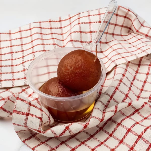 Two gulab jamuns in syrup inside a plastic container, with a plastic spoon, on a red and white checkered cloth.